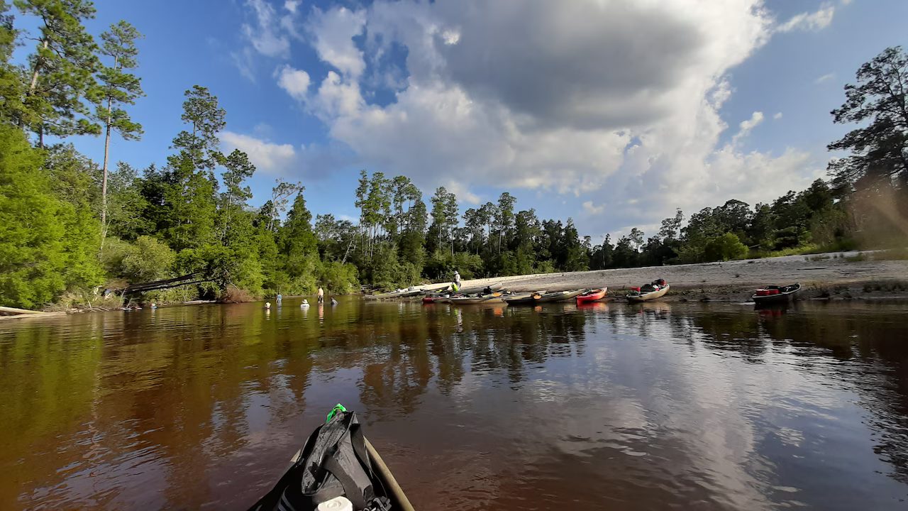 Kayak bow approaching a sandbar lined with beached canoes and kayaks
